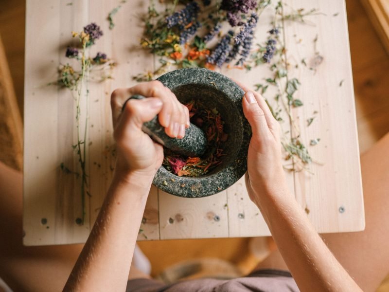 Top view of hands grinding dried herbs and petals with a mortar and pestle on a wooden table.